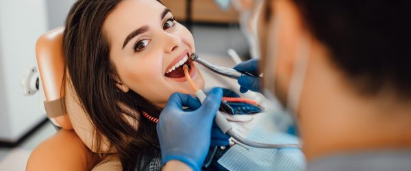 Young female patient visiting dentist office. Beautiful woman with healthy straight white teeth sitting at dental chair with open mouth during oral checkup while doctor working at teeth.